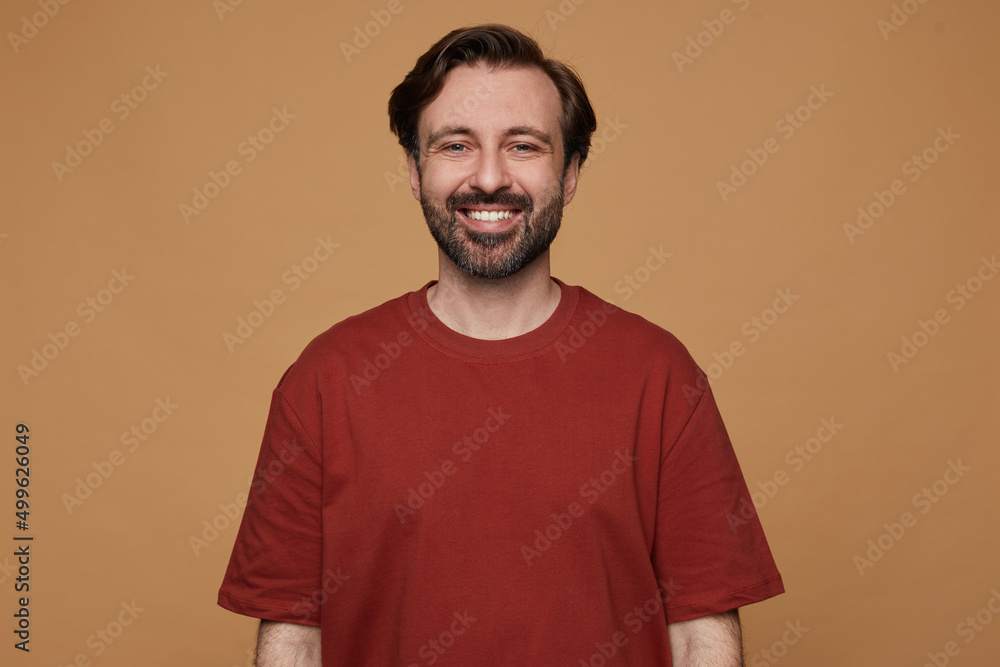indoor portrait of bearded male posing over beige background wears red t-shirt smiles and looks into camera with positive facial expression indoor portrait of bearded male posing over beige background wears red t-shirt smiles and looks into camera with positive facial expression