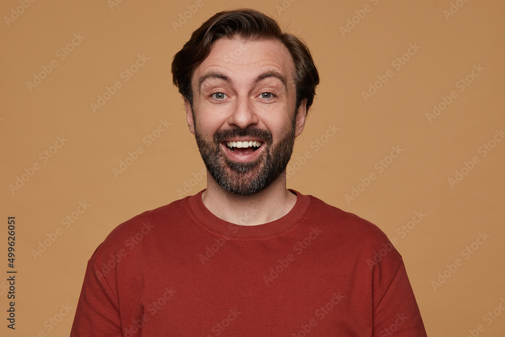 indoor portrait of bearded male posing over beige background wears red t-shirt smiles and looks into camera with positive surprised facial expression indoor portrait of bearded male posing over beige background wears red t-shirt smiles and looks into camera with positive surprised facial expression