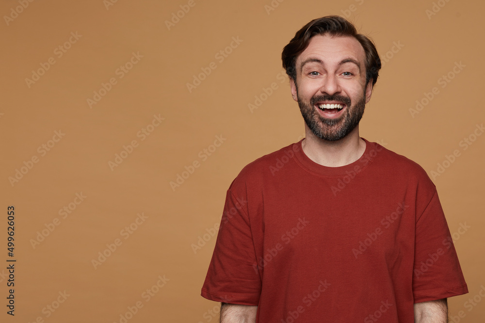 indoor portrait of bearded male posing over beige background wears red t-shirt smiles and looks into camera with positive surprised facial expression indoor portrait of bearded male posing over beige background wears red t-shirt smiles and looks into camera with positive surprised facial expression