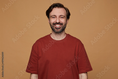 indoor portrait of bearded male posing over beige background wears red t-shirt smiles and looks into camera with positive facial expression
