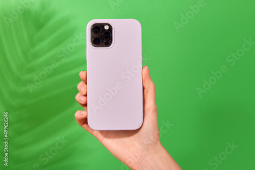 Female hand holding white smartphone in soft silicone cover back view . Phone case mock up isolated on green background shadow from a plant