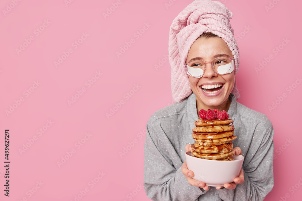 Young positive woman holds bowl with pile of delicious pancakes smiles toothily concentrated away applies beauty patches to reduce wrinkles feels refreshed after sleeping wears pajama stands indoor Young positive woman holds bowl with pile of delicious pancakes smiles toothily concentrated away applies beauty patches to reduce wrinkles feels refreshed after sleeping wears pajama stands indoor