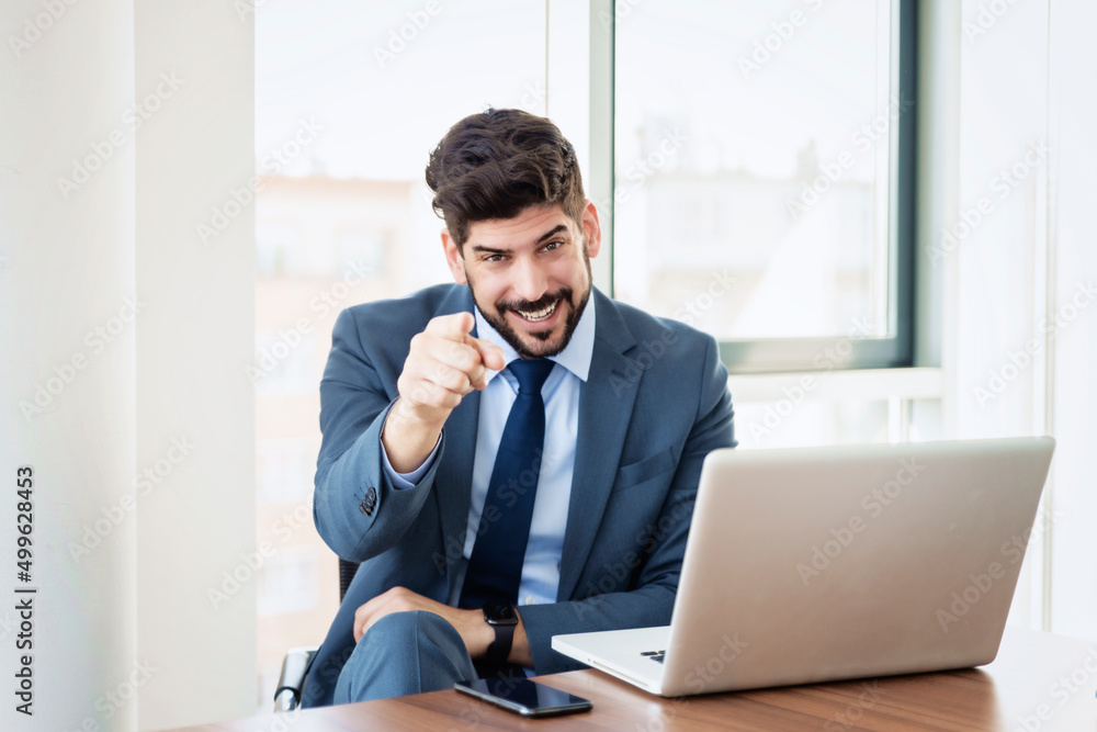 Confident businessman sitting behind his laptop and pointing at camera Confident businessman sitting behind his laptop and pointing at camera
