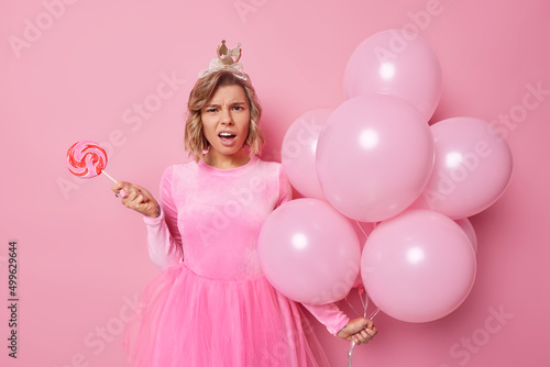 Pretty indignant woman holds caramel candy and bunch of balloons looks confused feels disappointed wears festive dress cannot understand why party is cancelled isolated over pink background. © wayhome.studio 