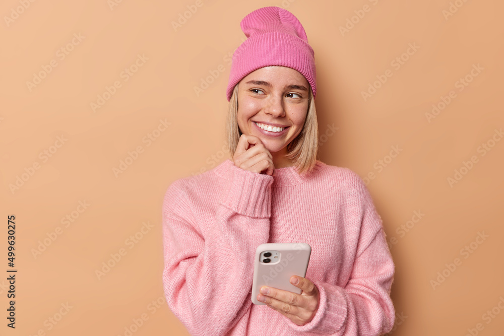 Pretty delighted woman looks awa with dreamy expression uses cellphone chats with friend posts something in social media wears pink jumper and hat poses over brown background thinks about mobile offer - obrazy, fototapety, plakaty 