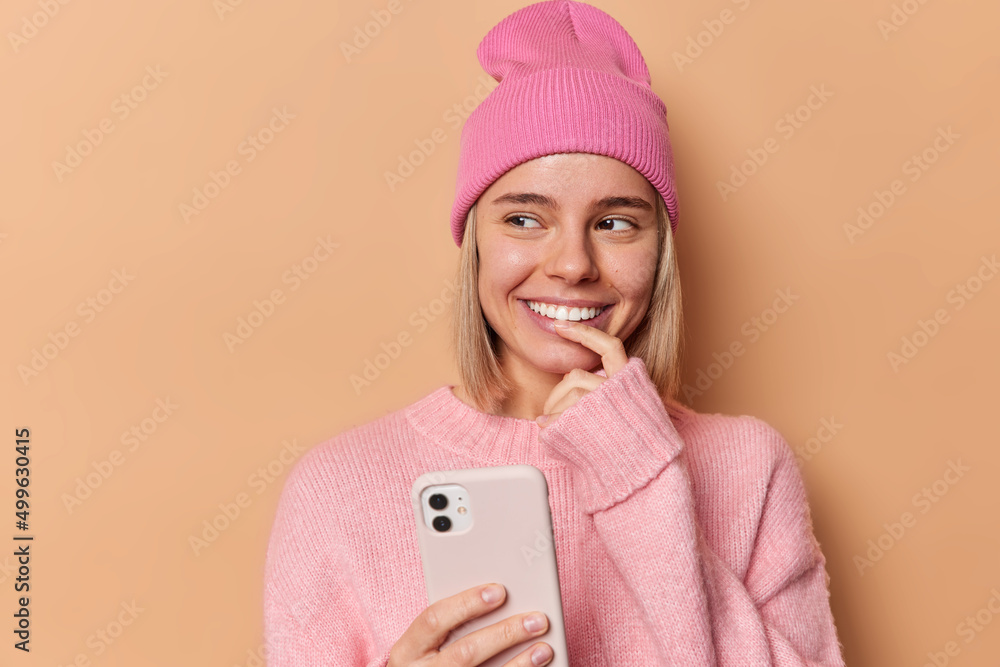 Lovely pensive European woman uses mobile phone thinks about message content scrolls social networks looks gladfully away wears soft loose pink jumper and hat isolated over brown background. Lovely pensive European woman uses mobile phone thinks about message content scrolls social networks looks gladfully away wears soft loose pink jumper and hat isolated over brown background.