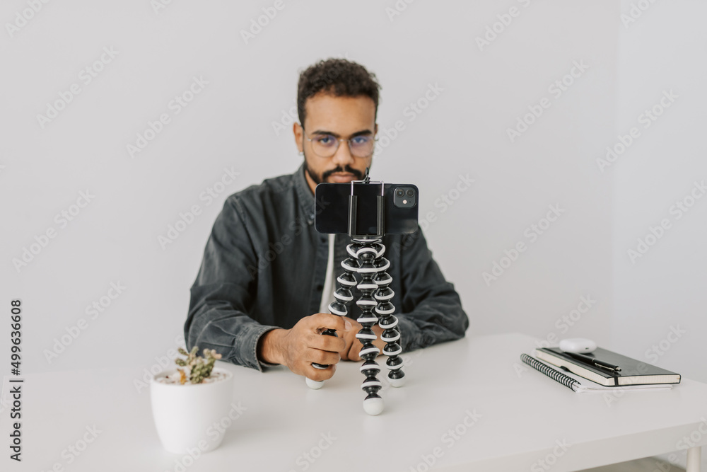 Young attractive afro-american man in grey shirt,eyeglasses sit at the desk, check social feed on black phone with tripod, touch screen, record videos close up. Live, camera ready for online teaching. Young attractive afro-american man in grey shirt,eyeglasses sit at the desk, check social feed on black phone with tripod, touch screen, record videos close up. Live, camera ready for online teaching.
