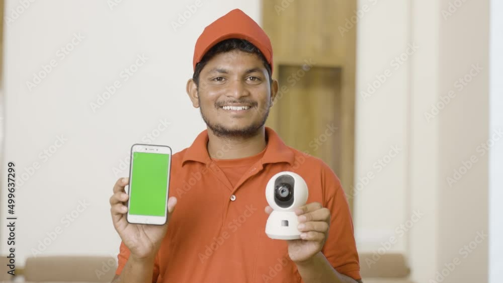 happy technician showing green screen mobile phone by holding smart cctv camera while looking camera at home - concept of technology, advertisement and professional occupation happy technician showing green screen mobile phone by holding smart cctv camera while looking camera at home - concept of technology, advertisement and professional occupation