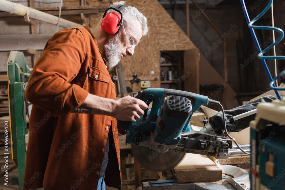 grey haired carpenter working with miter saw in woodwork studio.