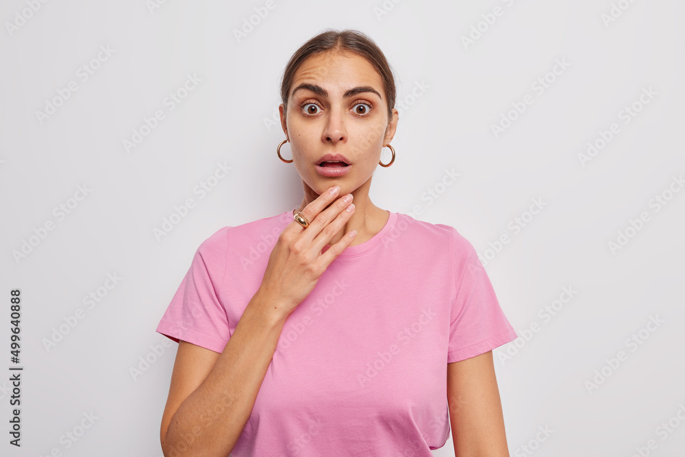 Indoor shot of amazed brunette European woman reacts on shocking news keeps hand on chin hears surprising terrible news wears casual pink t shirt and earrings isolated over white background. Indoor shot of amazed brunette European woman reacts on shocking news keeps hand on chin hears surprising terrible news wears casual pink t shirt and earrings isolated over white background.