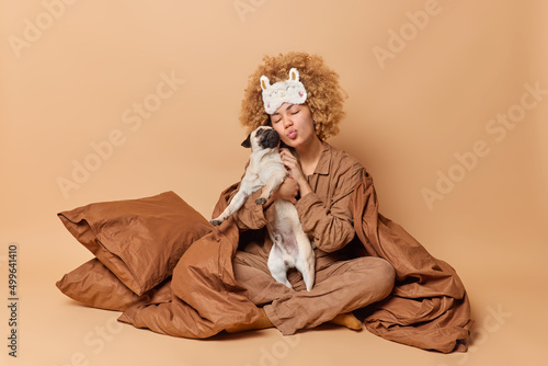 Caring woman takes care of her pet embraces pug dog with love keeps lips folded stays in bed under soft blanket enjoys time for rest isolated over brown background. Domestic animals concept.