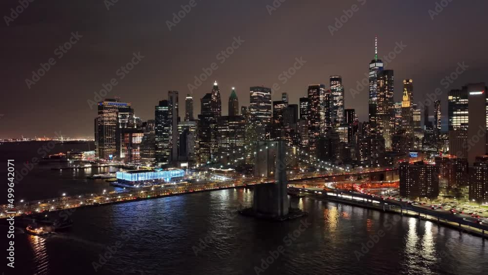 aerial across Manhattan skyline and Brooklyn Bridge American flag at night buildings skyscrapers lights on water NYC aerial across Manhattan skyline and Brooklyn Bridge American flag at night buildings skyscrapers lights on water NYC