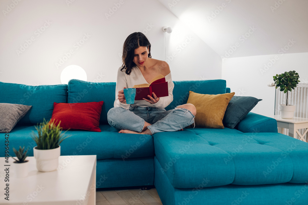 Woman reading a book and drinking tea while relaxing on a sofa at home Woman reading a book and drinking tea while relaxing on a sofa at home