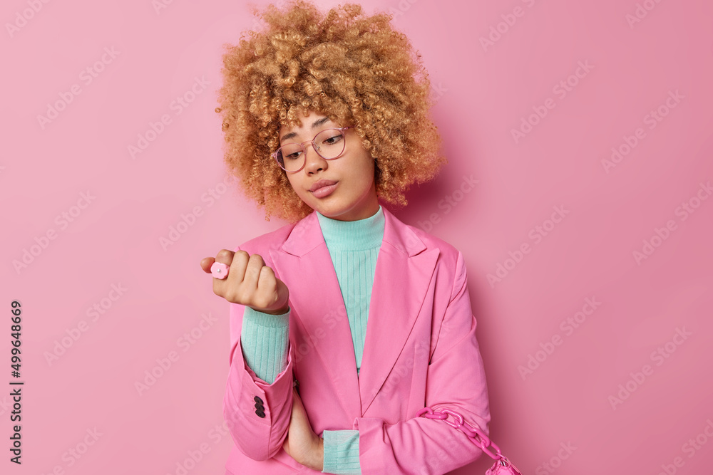 Serious curly haired young woman looks attentively at her nails feels bored while waiting for something dressed in elegant clothes isolated over pink background thinks about making new manicure