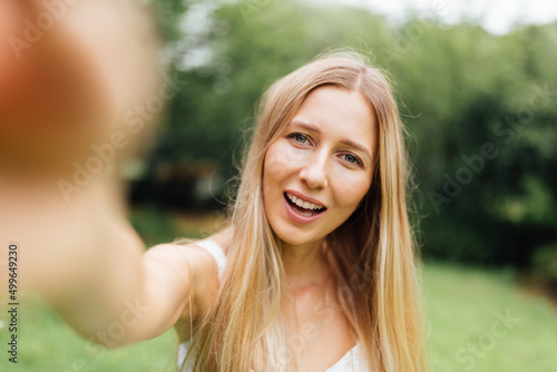 Portrait of beautiful blonde millennial woman making selfie outdoor. Young woman walking alone in the summer park after covid-19 coronavirus pandemic quarantine. Health care and happiness concept.