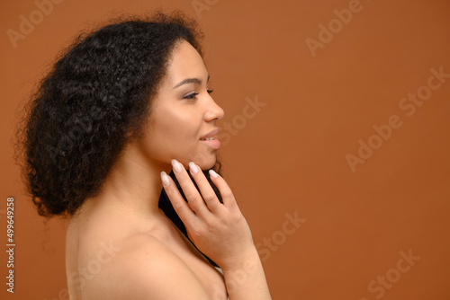 Side view at sensual naked beautiful african-american woman is touching gently face. Body and face care concept. Charming multiracial lady stands in profile isolated on brown background