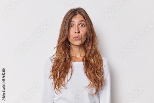 Facial expressions concept. Beautiful young woman with long wavy hair pouts lips wants to kiss someone looks surprisingly at camera dressed in casual sweater isolated over white studio background.