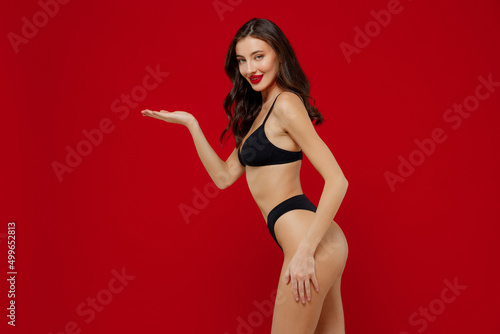 Profile young sexy woman 20s with perfect fit body in black underwear hold hand empty palm with workspace area mock up isolated on plain red background studio portrait. People female beauty concept
