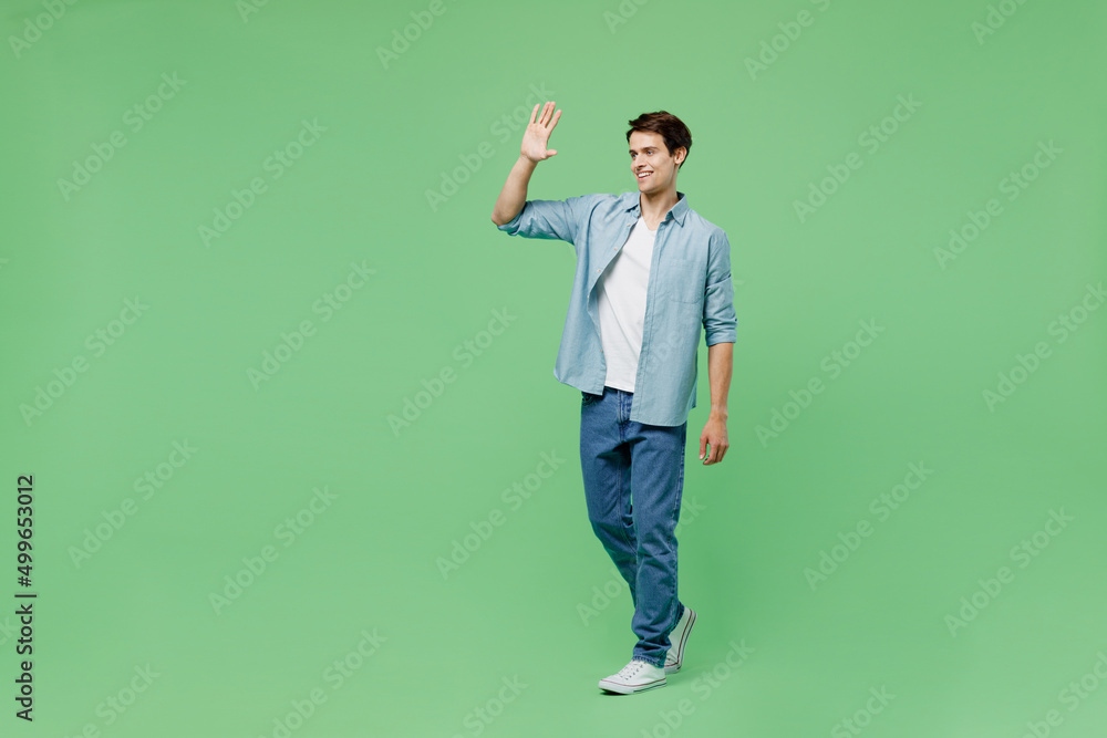 Full size body length side view cheerful happy fun smiling young brunet man 20s years old wears blue shirt meet greet waving hand as notices someone isolated on plain green background studio portrait. Full size body length side view cheerful happy fun smiling young brunet man 20s years old wears blue shirt meet greet waving hand as notices someone isolated on plain green background studio portrait.