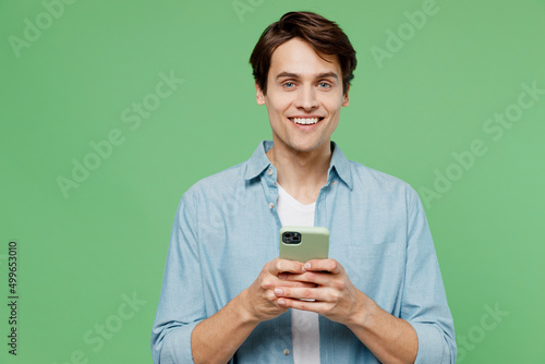 Overjoyed excited jubilant happy young brunet man 20s years old wear blue shirt hold in hand use mobile cell phone typing browsing chatting send sms isolated on plain green background studio portrait