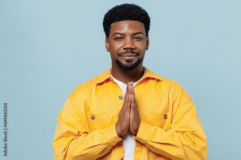 Obraz premium Young man of African American ethnicity 20s in yellow shirt hold hands folded in prayer gesture, begging about something isolated on plain pastel light blue background studio People lifestyle concept