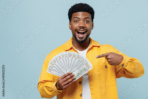 Young shocked amazed fun man of African American ethnicity 20s wear yellow shirt hold point index finger on fan of cash money in dollar banknotes isolated on plain pastel light blue background studio photo