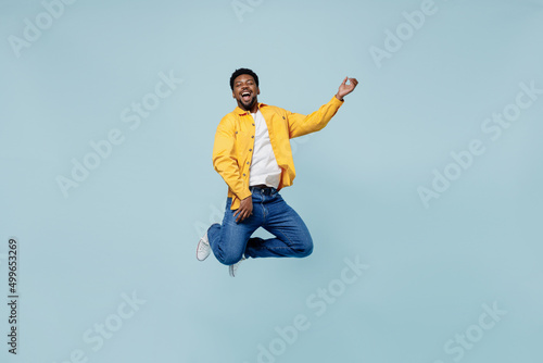 Full body expressive singer young man of African American ethnicity 20s in yellow shirt jump high play guitar isolated on plain pastel light blue background studio portrait. People lifestyle concept