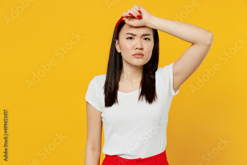 Sick ill tired frowning young girl woman of Asian ethnicity 20s years old wears white t-shirt hand on forehead isolated on plain yellow background studio portrait. People emotions lifestyle concept.