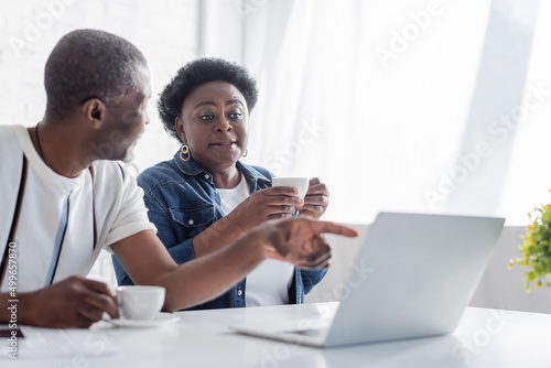 retired african american man pointing at laptop near wife with cup.