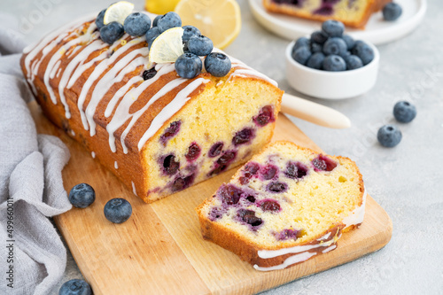 Lemon blueberry cake with lemon icing and fresh berries on top on the board on a gray concrete background with cup of tea. Selective focus. Copy space.