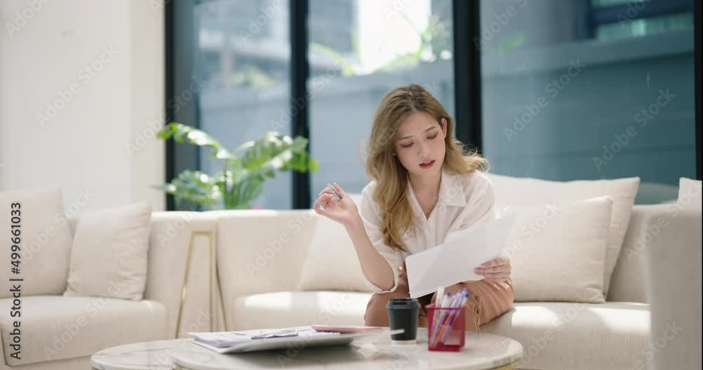 Young Asian woman sitting on couch holds paper receipt calculates monthly expenses using calculator check utility bills managing family budget, accountant do paperwork job at home concept. Young Asian woman sitting on couch holds paper receipt calculates monthly expenses using calculator check utility bills managing family budget, accountant do paperwork job at home concept.