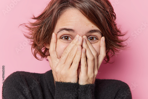 Foto Portrait of happy brunette woman with bob hairstyle tries to hide keeps hands on face has eyes full of happiness feels very glad dressed in casual black jumper poses against pink background