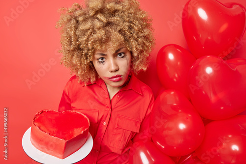 Serious woman with spoiled makeup after crying wears shirt holds heart shaped sweet cake and bunch of balloons prepares for holiday celebration isolated over vivid red backgroud. Bad day concept