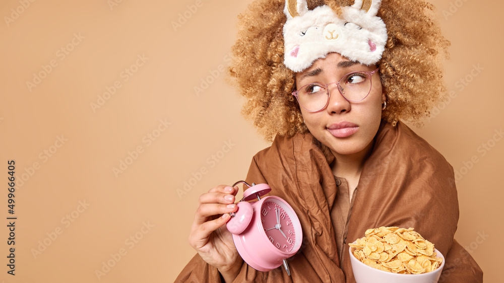 Displeased young woman wakes uo early in morning going to have breakfast holds alarm clock and bowl of cornflakes concentrated away poses against brown background mock up space for your text Displeased young woman wakes uo early in morning going to have breakfast holds alarm clock and bowl of cornflakes concentrated away poses against brown background mock up space for your text