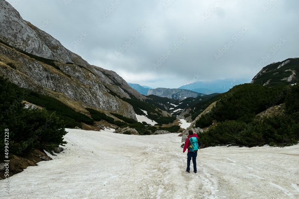 Woman with backpack crossing a snow field on a hiking trail with view on cloud covered mountain peaks of the Hochschwab Region in Upper Styria, Austria. Plateau full of snow in beautiful Alps, Europe.