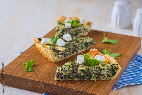 Unsweetened sliced open pie, shortcrust pastry quiche stuffed with spinach, mushrooms and cheese on a wooden board on a light concrete background. Unsweetened pastry recipes.