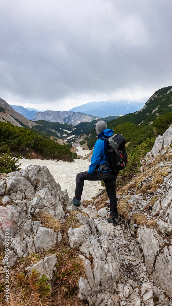 Man standing on rock with backpack on scenic hiking trail with view on cloud covered mountain peaks of the Hochschwab Region in Upper Styria, Austria. Snow covered meadows in beautiful Alps in Europe