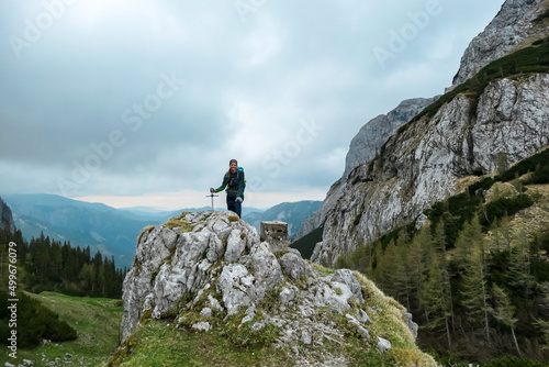 Woman with backpack standing on rock next to the Haeuslalm cottage. Hiking trail in early spring with panoramic view on the mountain peaks of Hochschwab Region in Upper Styria, Austria. Alps, Europe