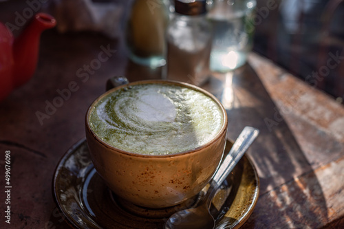 Green Matcha Latte, popular trendy tea with latte art heart shape on top. Served in cafe in ceramic mug on rustic table. Strong shadows and summer sun light
