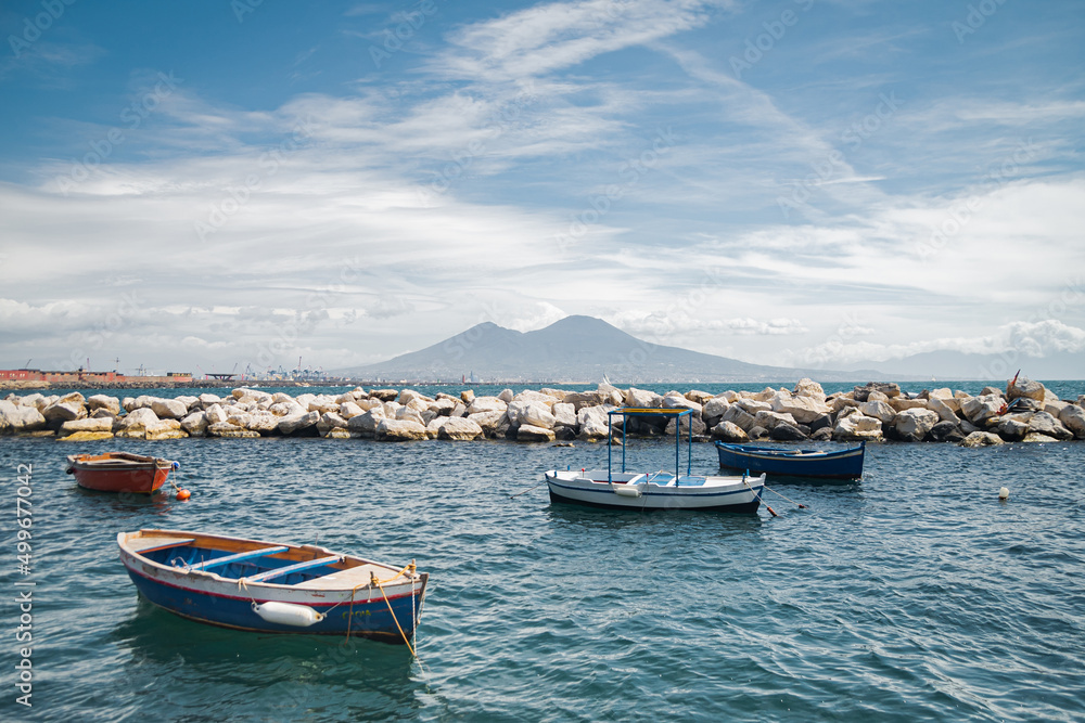 Calm blue Tyrrhenian Sea. View from the embankment of Naples to Mount Vesuvius volcano. Pleasure boats moored near the shore and stone breakwaters.