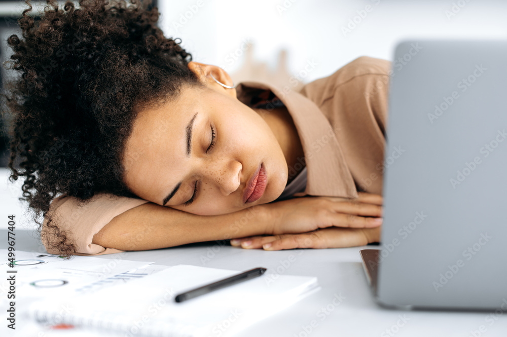 Close-up of a tired exhausted african american girl, female freelancer or student, fell asleep at the desktop near the laptop, overworked from working or studying online, needs rest and sleep Close-up of a tired exhausted african american girl, female freelancer or student, fell asleep at the desktop near the laptop, overworked from working or studying online, needs rest and sleep