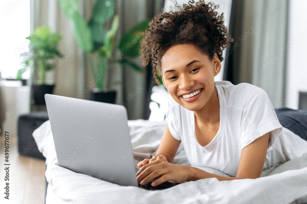 Lovely positive african american curly-haired brunette girl lies at home on a bed on stomach, uses a laptop, browses the Internet, chatting on social networks, looks at camera, smiles happily Lovely positive african american curly-haired brunette girl lies at home on a bed on stomach, uses a laptop, browses the Internet, chatting on social networks, looks at camera, smiles happily