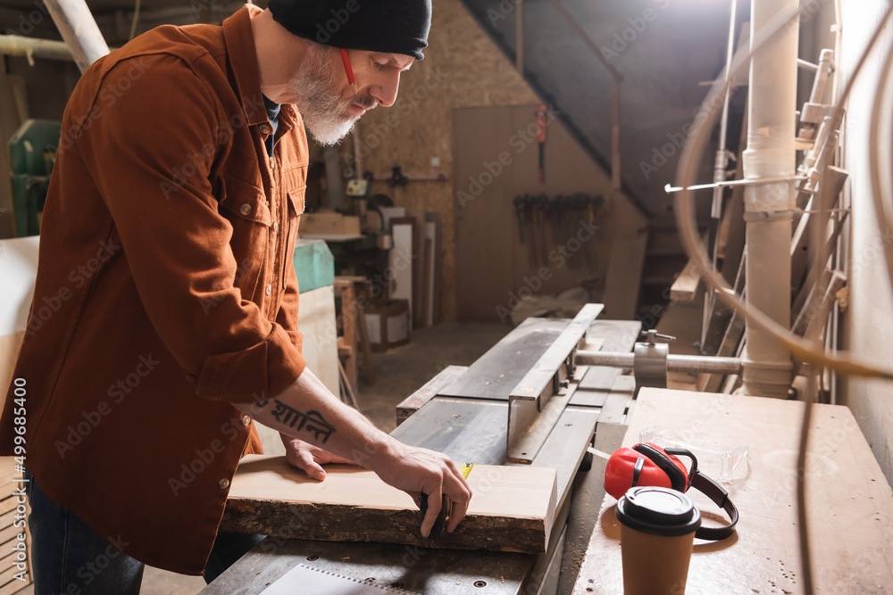 side view of carpenter in beanie measuring board near coffee to go.