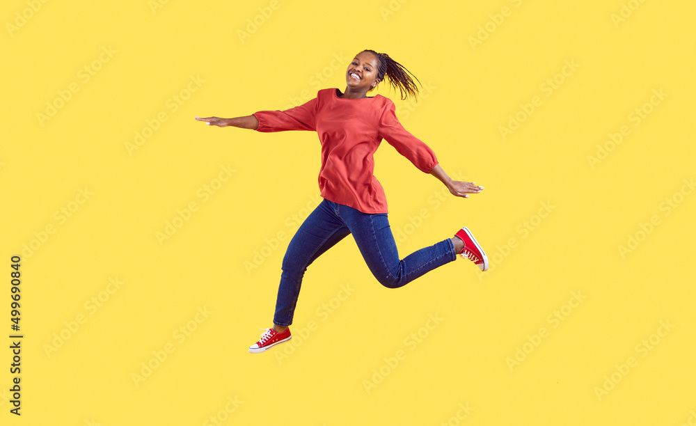 Happy black woman jumping in the studio. Positive carefree young Afro American girl wearing comfortable red blouse and blue jeans jumping high in air isolated on vibrant vivid yellow colour background