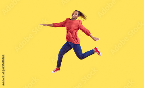 Happy black woman jumping in the studio. Positive carefree young Afro American girl wearing comfortable red blouse and blue jeans jumping high in air isolated on vibrant vivid yellow colour background