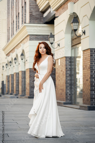 Refined feminine lady with red hair in a white dress. A happy and satisfied bride poses on a city street. Summer mood