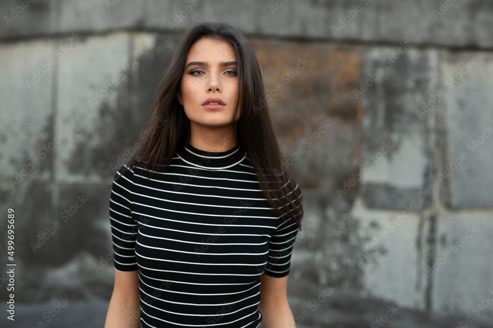 Young woman with straight brunette hair and makeup, looking sad at camera, over black old wall background.