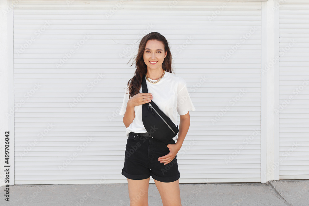 Woman, wearing white t-shirt, black shorts, fanny pack or waist pack and gold chain necklace, standing outdoor near white wall. Stylish trendy basic minimalistic casual outfit. Street fashion. Woman, wearing white t-shirt, black shorts, fanny pack or waist pack and gold chain necklace, standing outdoor near white wall. Stylish trendy basic minimalistic casual outfit. Street fashion.