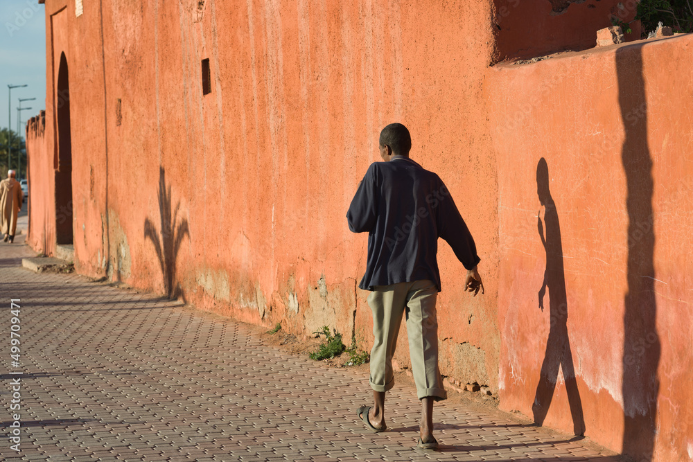 A pedestrian and his shadow walking in the street along an orange wall in Marrakech