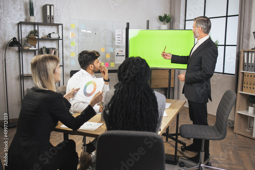 Successful mature business man presenting analytical report on green chroma key screen while working together with his concentrated multiracial colleagues in the modern office.